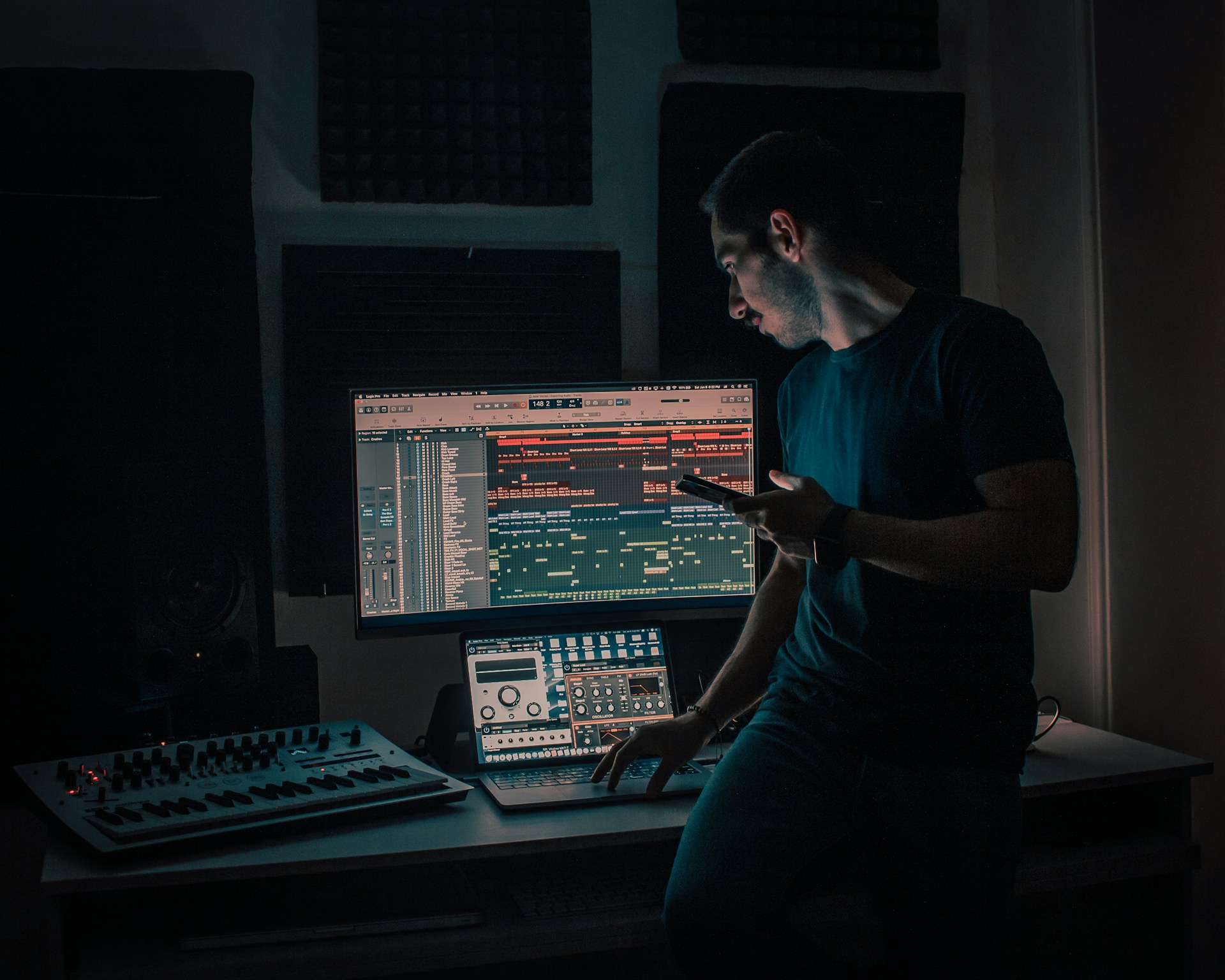 A producer working at a studio desk with a synthesiser, laptop and monitor showing a busy arrangement.